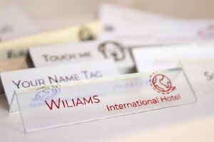 Clear plastic nameplate with the name “WILIAMS,” the phrase “International Hotel” and a small globe design printed in red on the front. An assortment of other silver and gold nameplates sit in the background, out of focus.