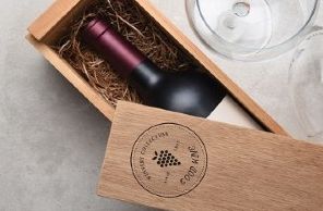 Bottle of red wine nested in a wooden box with two wine glasses beside it. The box has a custom logo on the lid with the words “Winery Collection Since 1845 — Good Wine.”